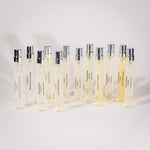 A set of 12 clear glass Tuckson Eau de parfum sample vials with silver caps, arranged in two rows on a light gray background. The vials contain a pale yellow liquid and are labeled with various fragrance names including 'Tuckson' and 'Monastic Flower'.