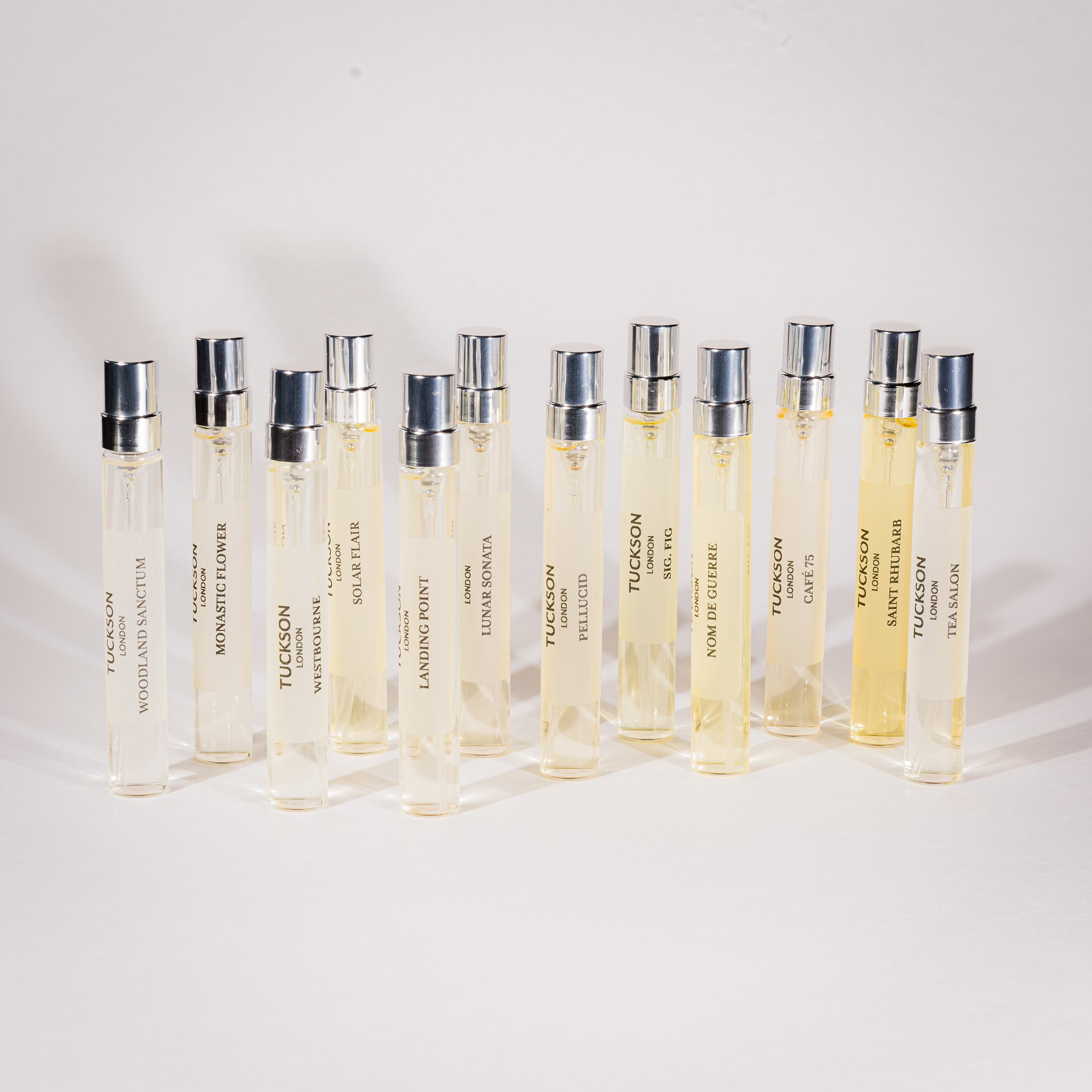 A set of 12 clear glass Tuckson Eau de parfum sample vials with silver caps, arranged in two rows on a light gray background. The vials contain a pale yellow liquid and are labeled with various fragrance names including 'Tuckson' and 'Monastic Flower'.