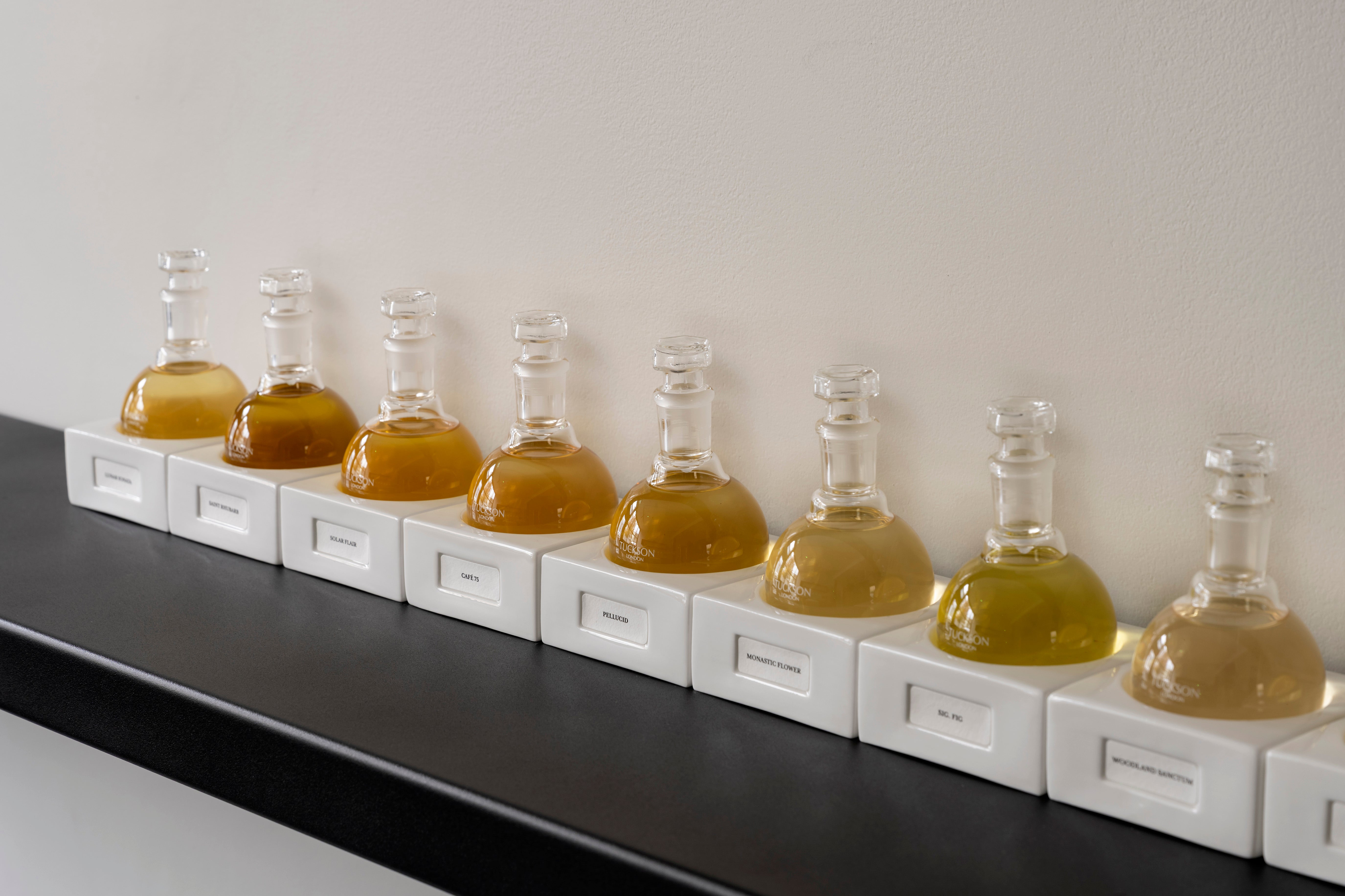 Row of perfume glass flasks with sitting on their respective porcelain bases on a black shelf against a white wall.
