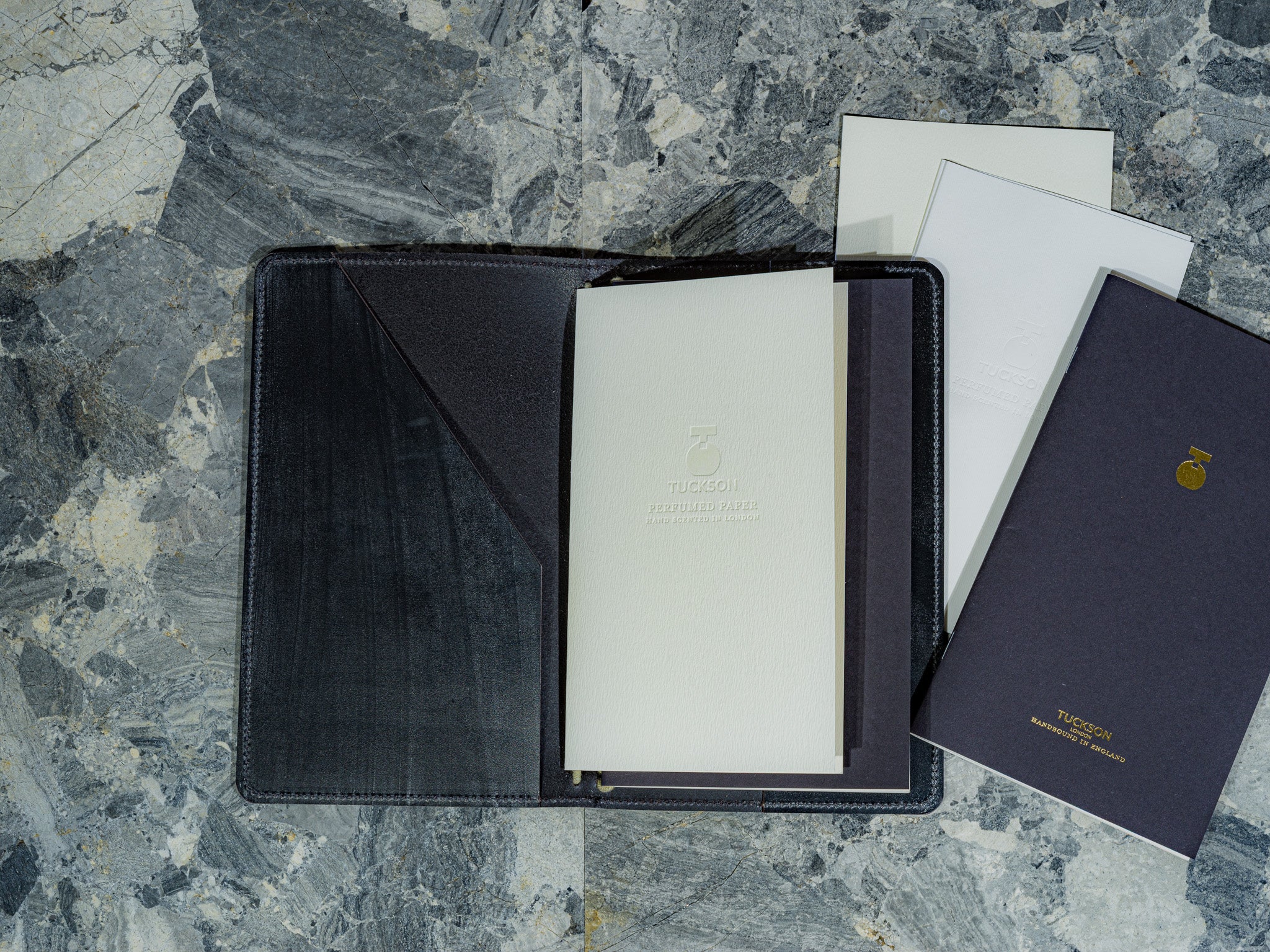 A black leather travel folio with a set of off white perfumed Tuckson cards, alongside a dark perfumed navy blue notebook with gold lettering, all arranged on a gray marble surface. 