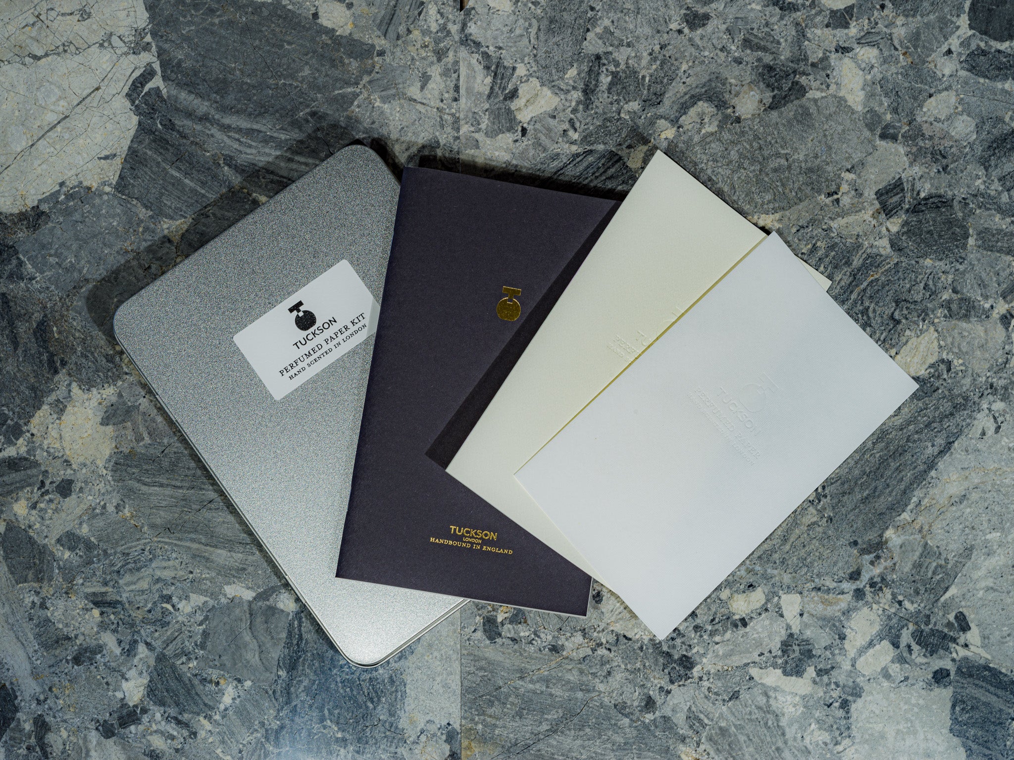 A set of three Tuckson Perfumed Paper stationery items arranged on a gray marbled surface: a silver tin storage case with a white brand sticker, a dark navy notebook with gold foil branding, and two off-white paper cards.